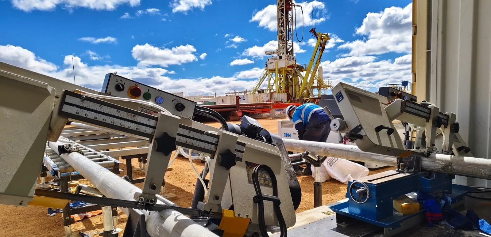 Two band saws cutting core on the processing tables at a rig site, the derrick behind