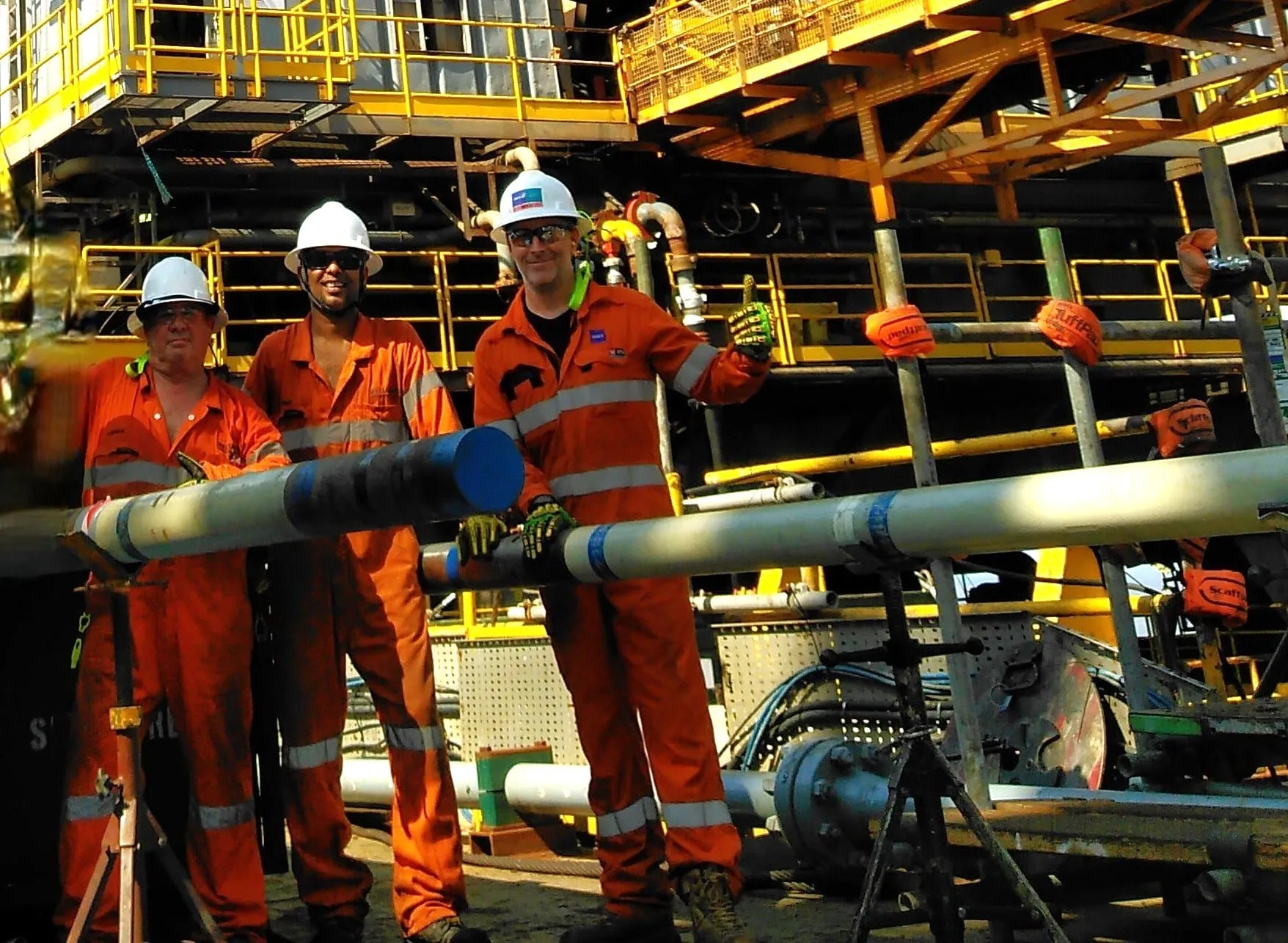 Three crew in orange coveralls on an offshore rig floor, holding a core barrel section